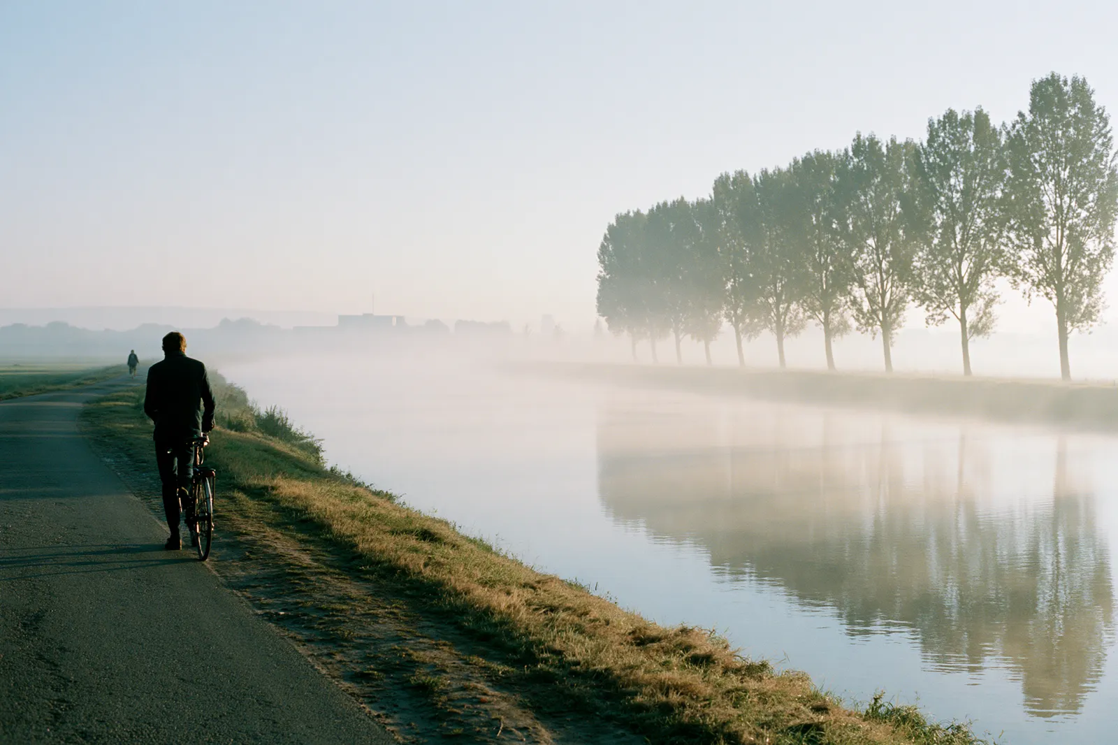 Homme marchant avec son vélo le long d'un canal belge au petit matin